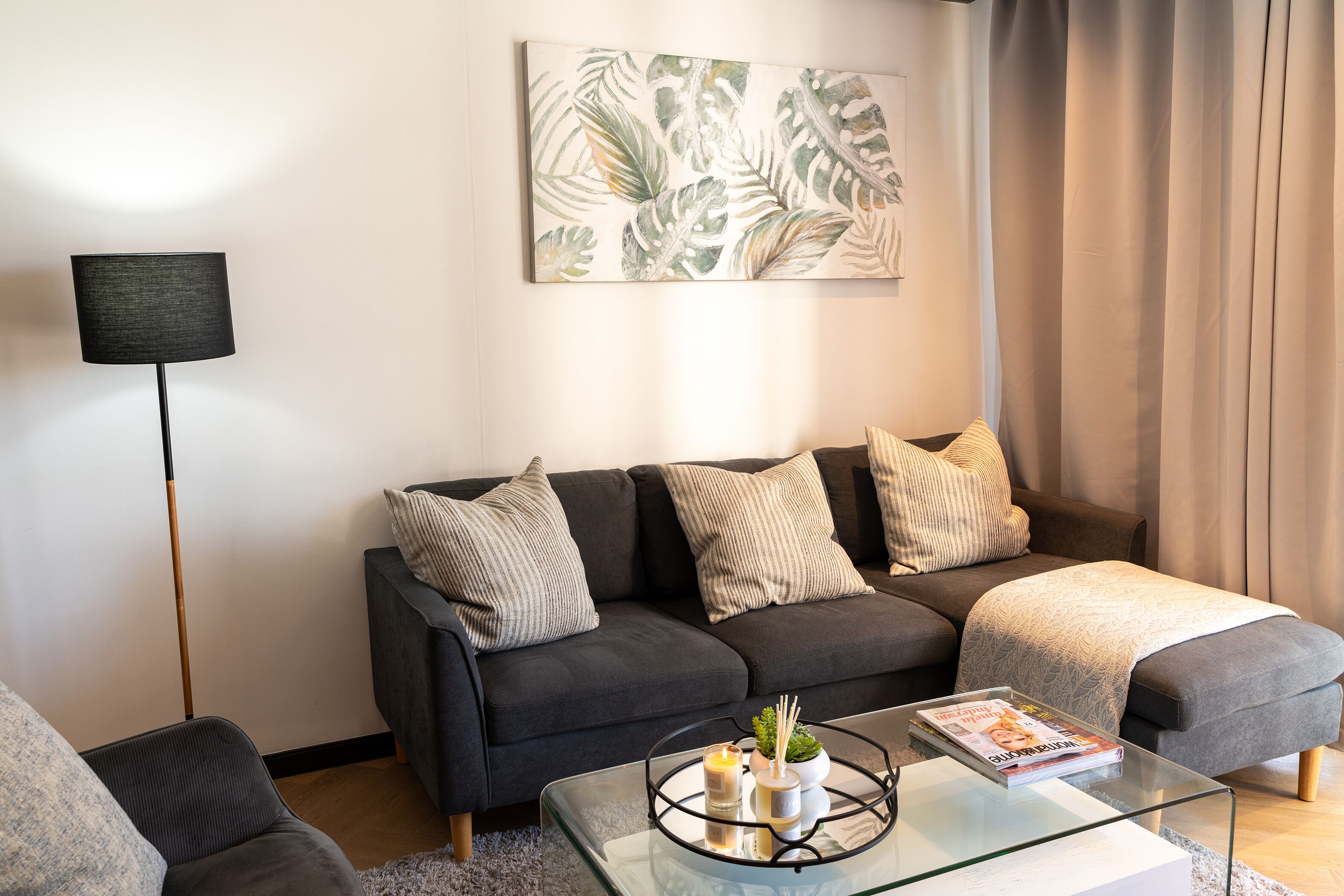 Ett modernt vardagsrum med en mörkgrå soffa, ett soffbord i glas och en golvlampa.