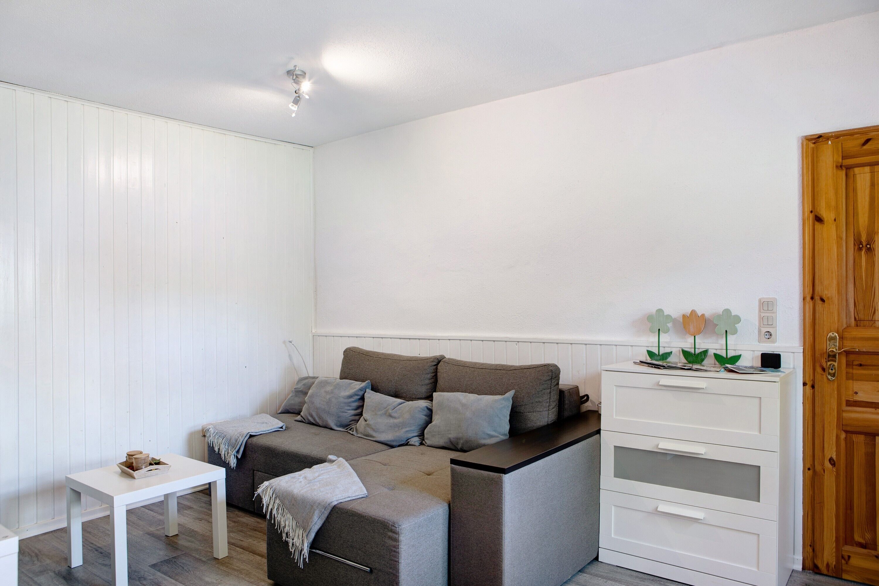 Een kamer met een grijze bank, een klein wit tafeltje, een witte kapstok en een houten deur.