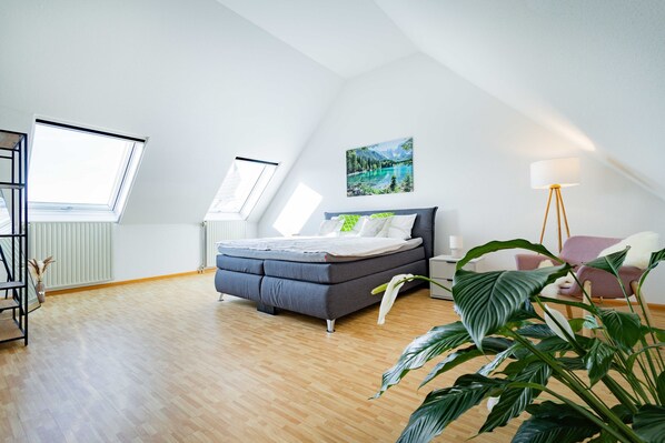 Une chambre à coucher avec un lit, des fenêtres de toit, un tableau, une lampe sur pied et une plante.