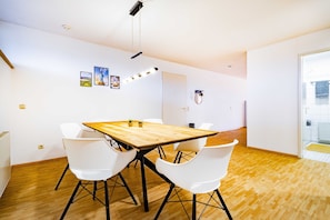 Une pièce moderne avec une table en bois, des chaises blanches, un luminaire au plafond et une porte donnant sur une salle de bain.