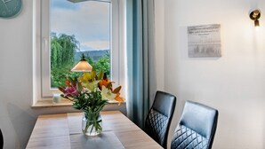 Uma área de jantar com mesa, cadeiras, um vaso de flores, relógio, janela com vista e luminária na parede.