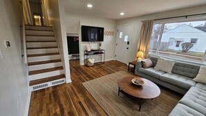 Una sala de estar con una escalera, un sofá, una mesa de centro, un televisor y una ventana con vistas a una casa.