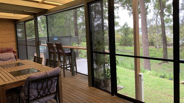 A spacious deck with a wooden table and chairs, large windows offering views of a garden and trees.