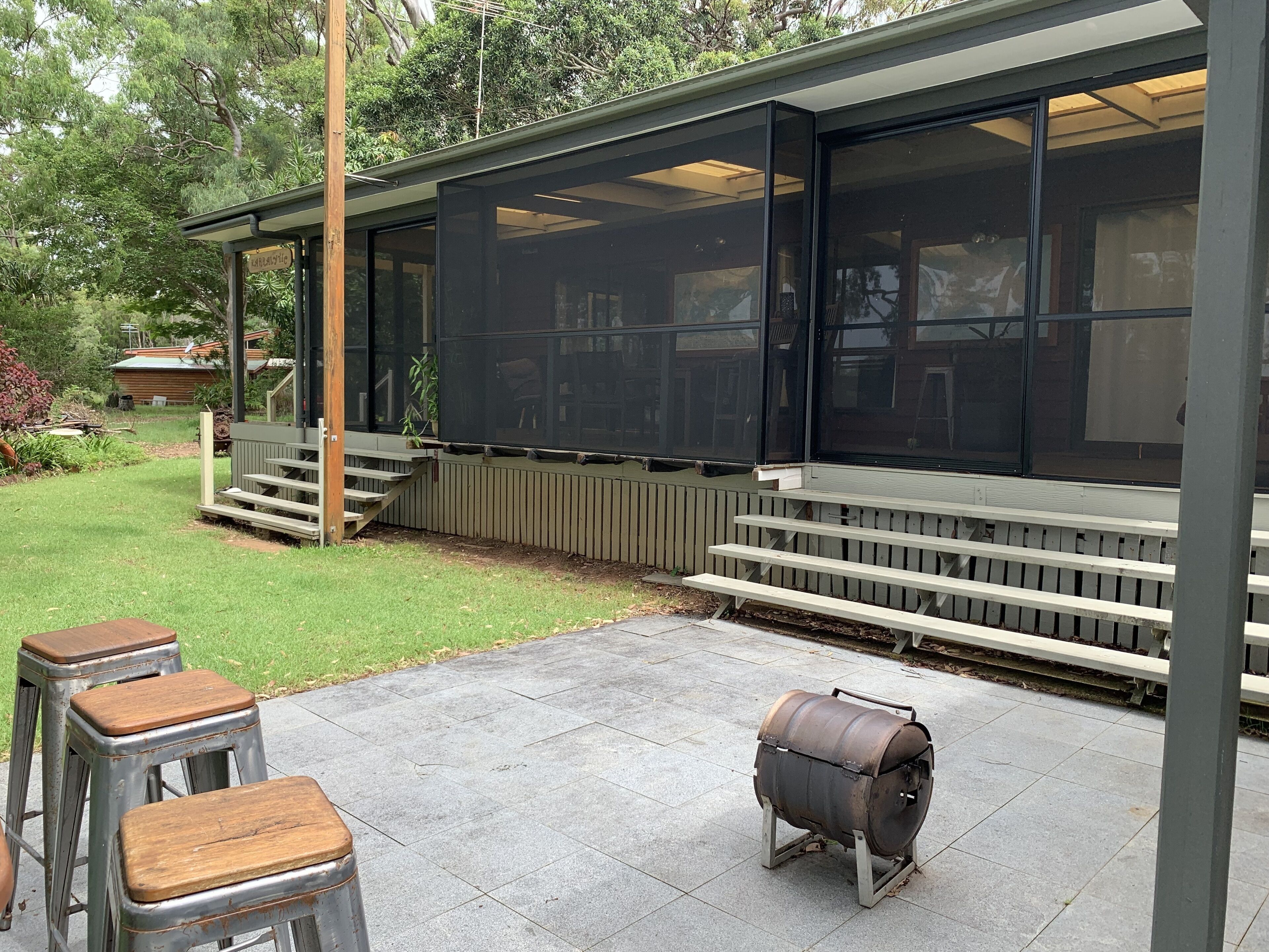 A covered outdoor patio with a barbecue grill, wooden stools, and a wooden deck with steps leading to a covered area.