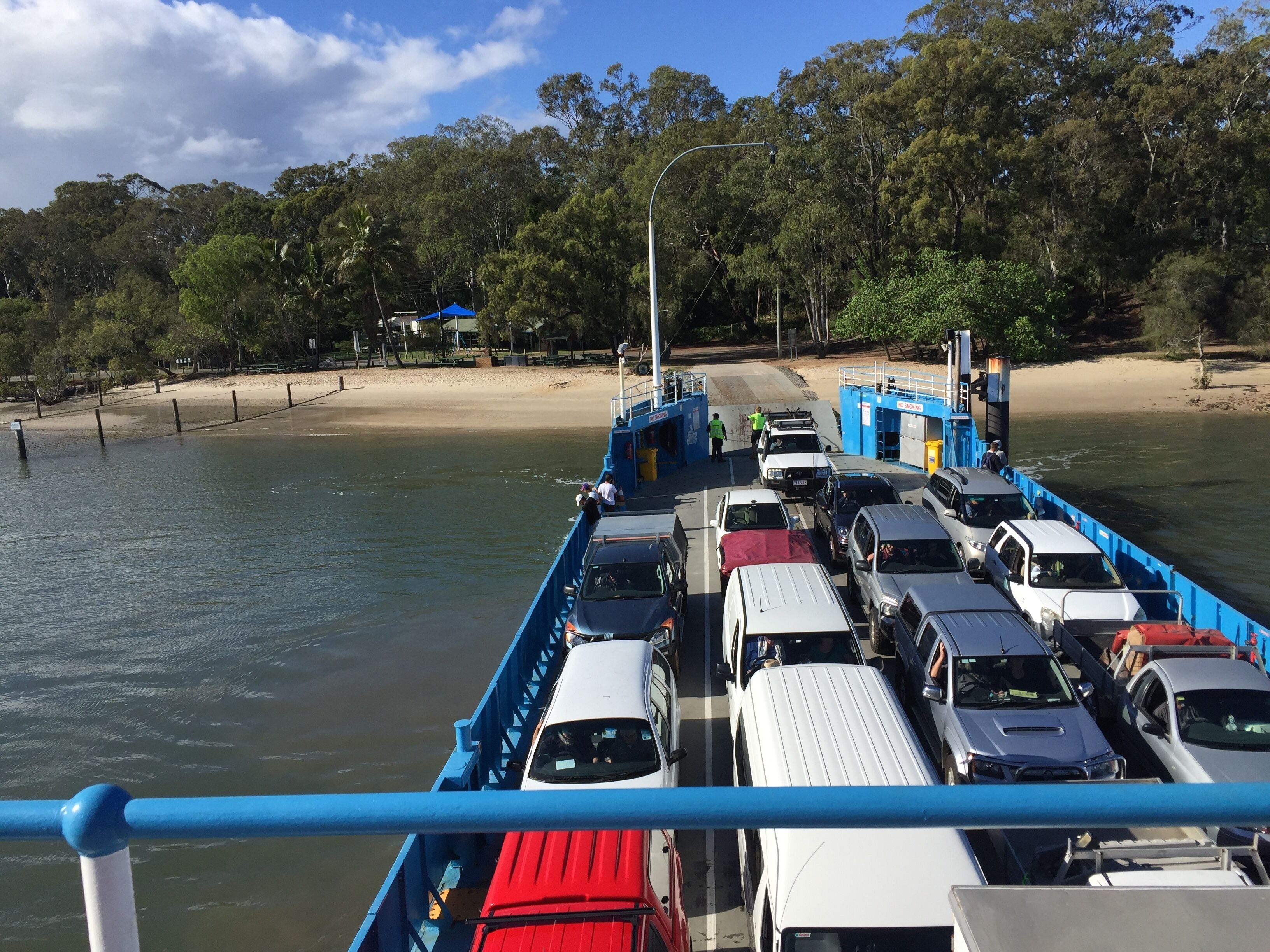 A ferry with multiple vehicles parked along the side, a beach, and trees in the background.
