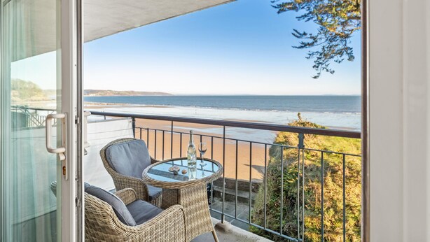Ein Balkon mit Korbmöbeln und einem Glastisch, mit Blick auf einen Strand und den Ozean.