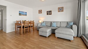 Ein Wohnzimmer mit grauem Sofa, Esstisch aus Holz und Stühlen. An der Wand hängen gerahmte Bilder und ein Fenster mit Vorhängen.