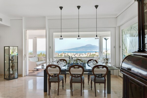 A dining area with a view of a city and mountains, featuring a glass table, wooden chairs, and pendant lights.