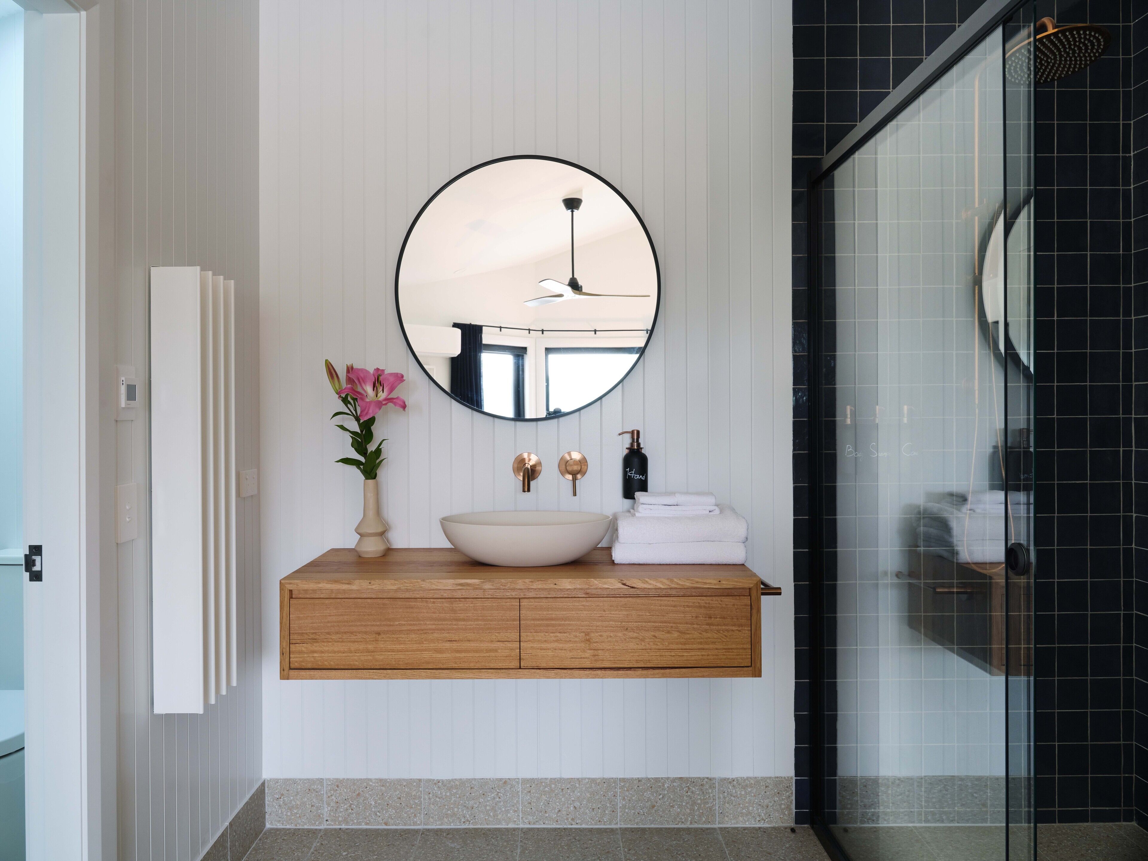 A bathroom with a round mirror, a white sink, a wooden vanity, a pink flower vase, and a glass shower enclosure.