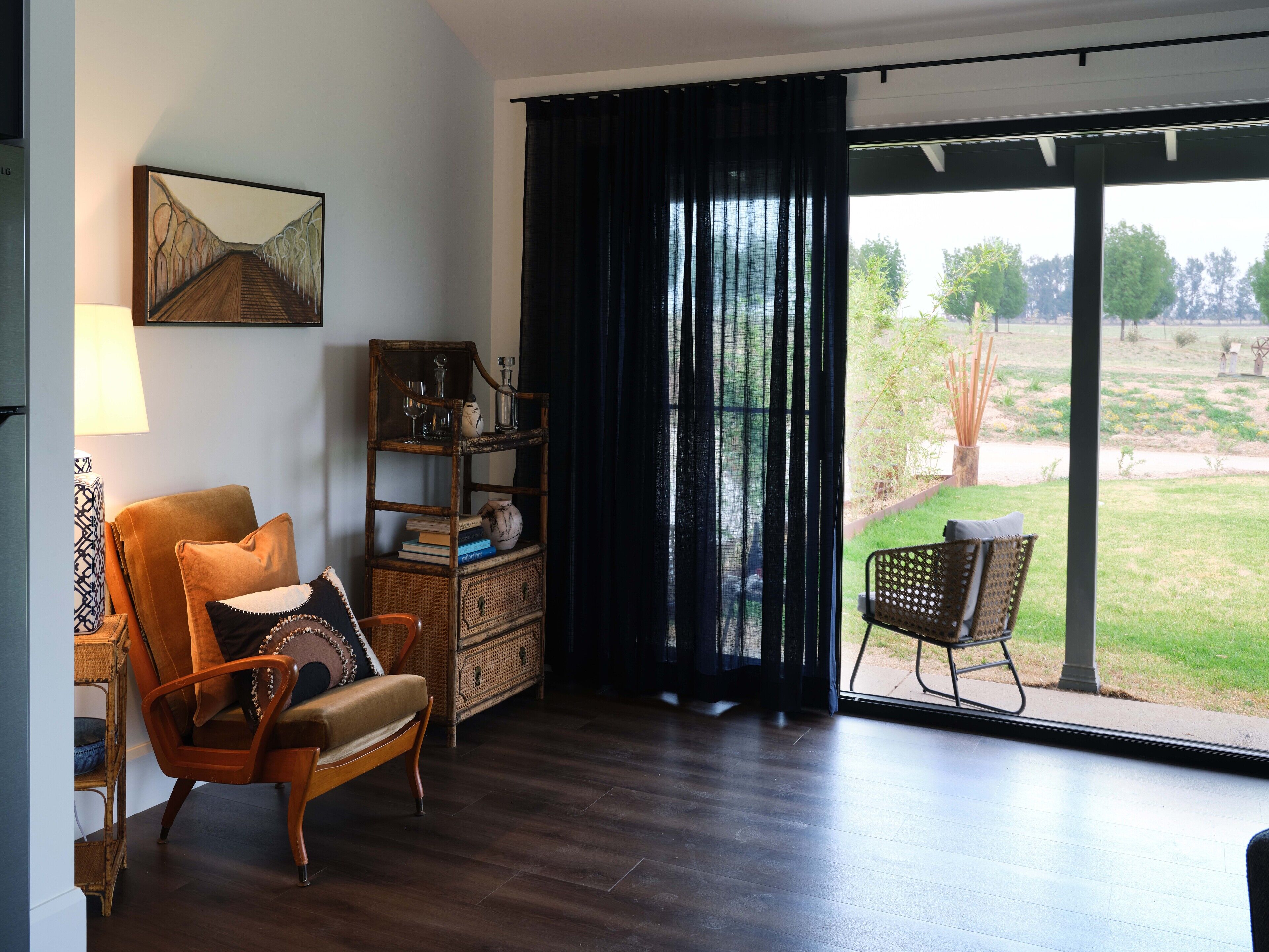 A room with a wooden floor, a brown leather chair, a wicker chair, a bookshelf, and a large window with a view of a garden.