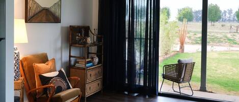 A room with a wooden floor, a brown leather chair, a wicker chair, a bookshelf, and a large window with a view of a garden.