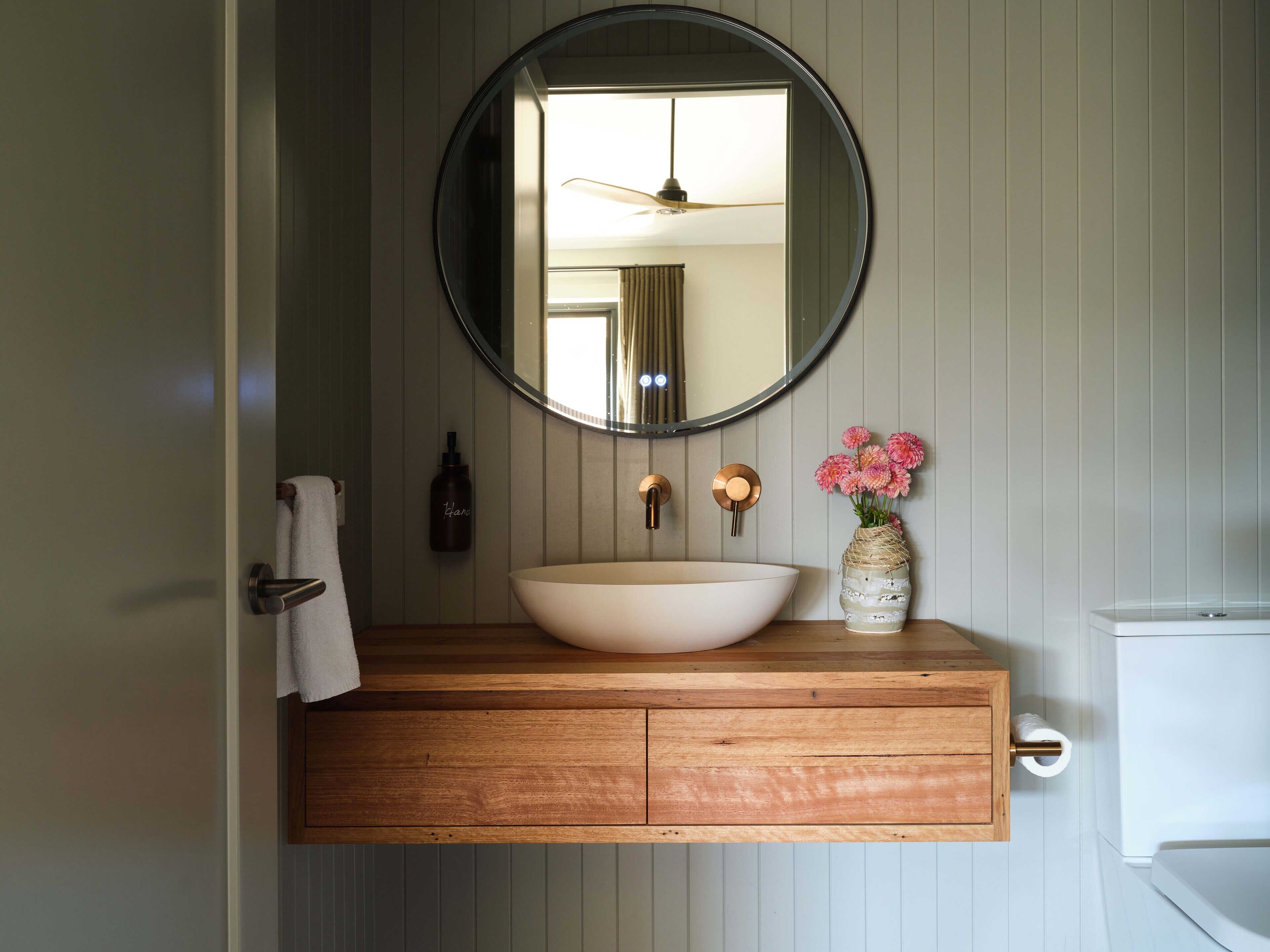 A bathroom with a wooden vanity, a round mirror, a white sink, a vase with pink flowers, and a towel hanging on the wall.