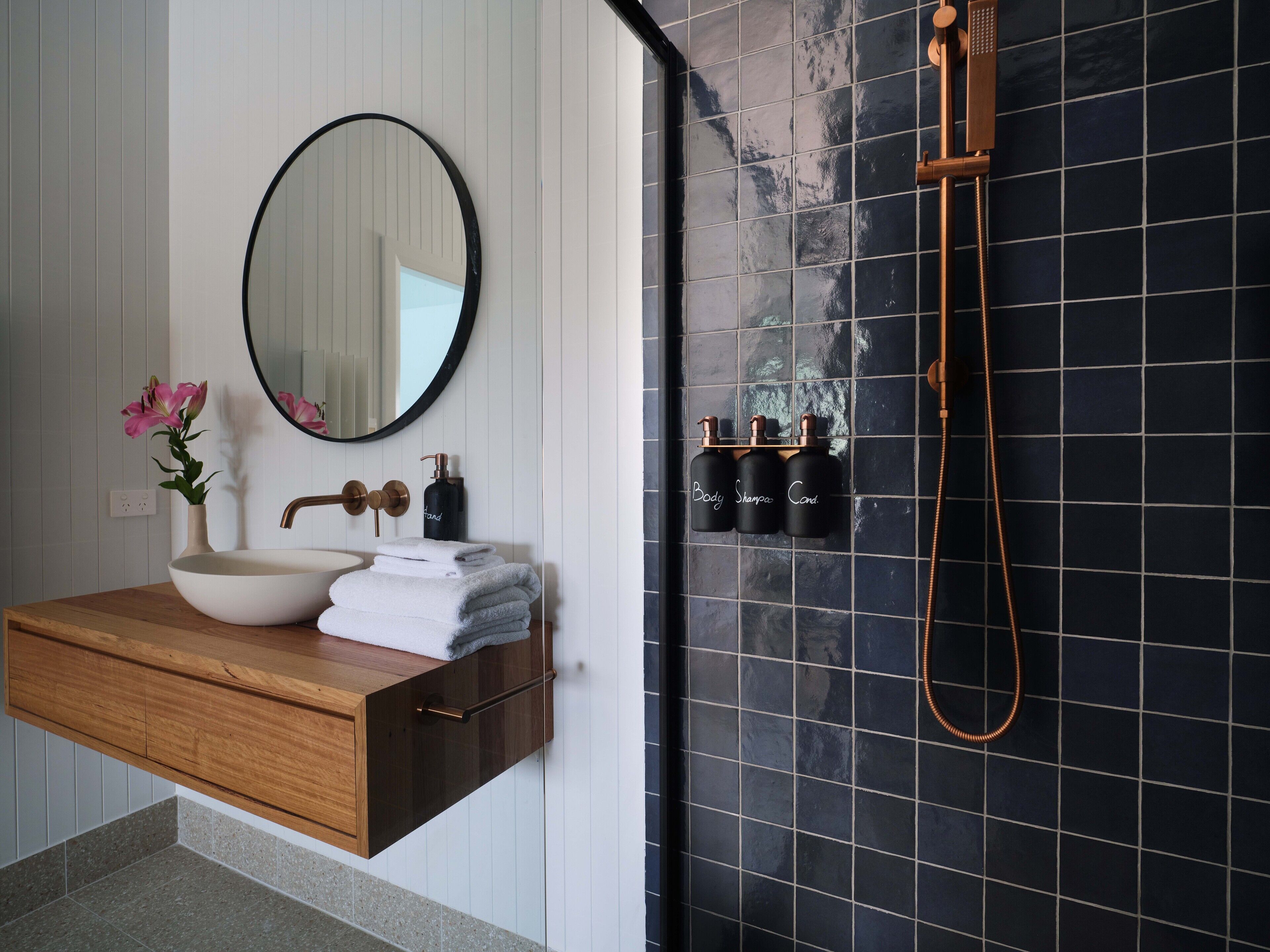 A bathroom with a wooden vanity, a round mirror, a white sink, a vase with pink flowers, a black bottle, and a black shower with a handheld showerhead.