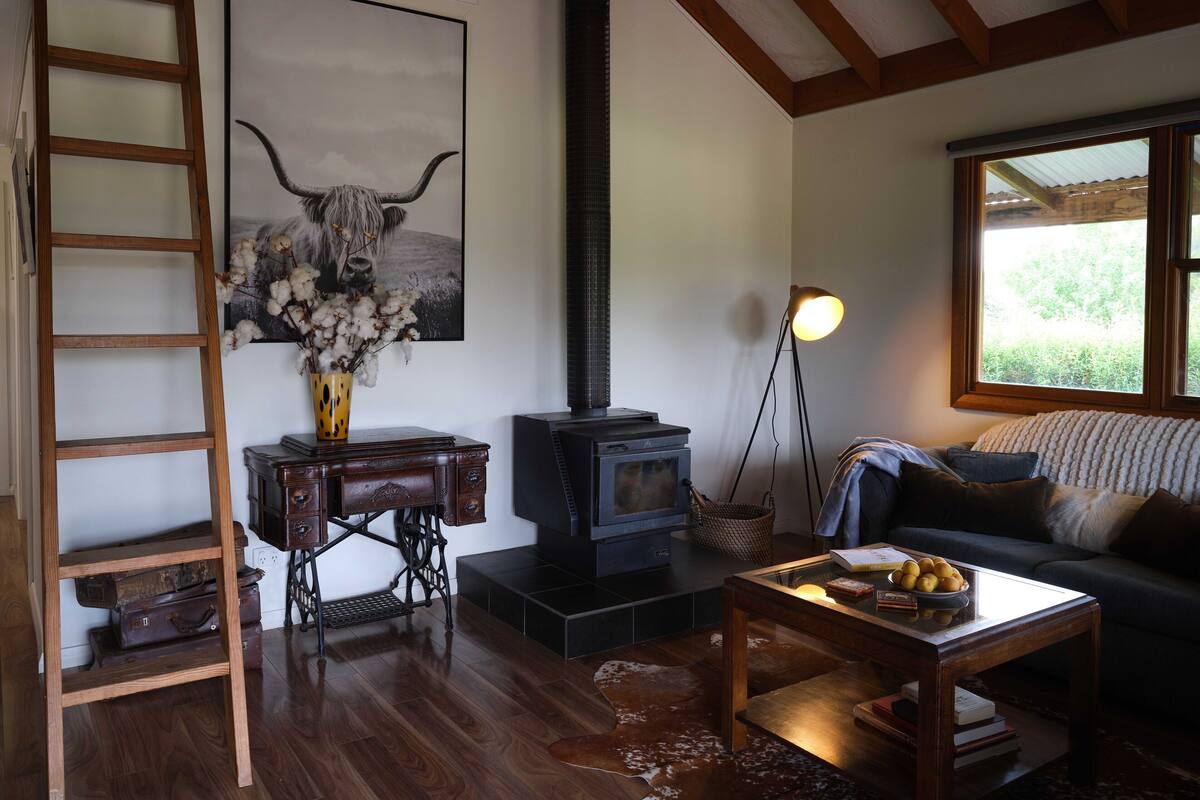 A cozy living room with a wooden floor, a sofa, a coffee table, a fireplace, a ladder, and a framed artwork of a cow's head.