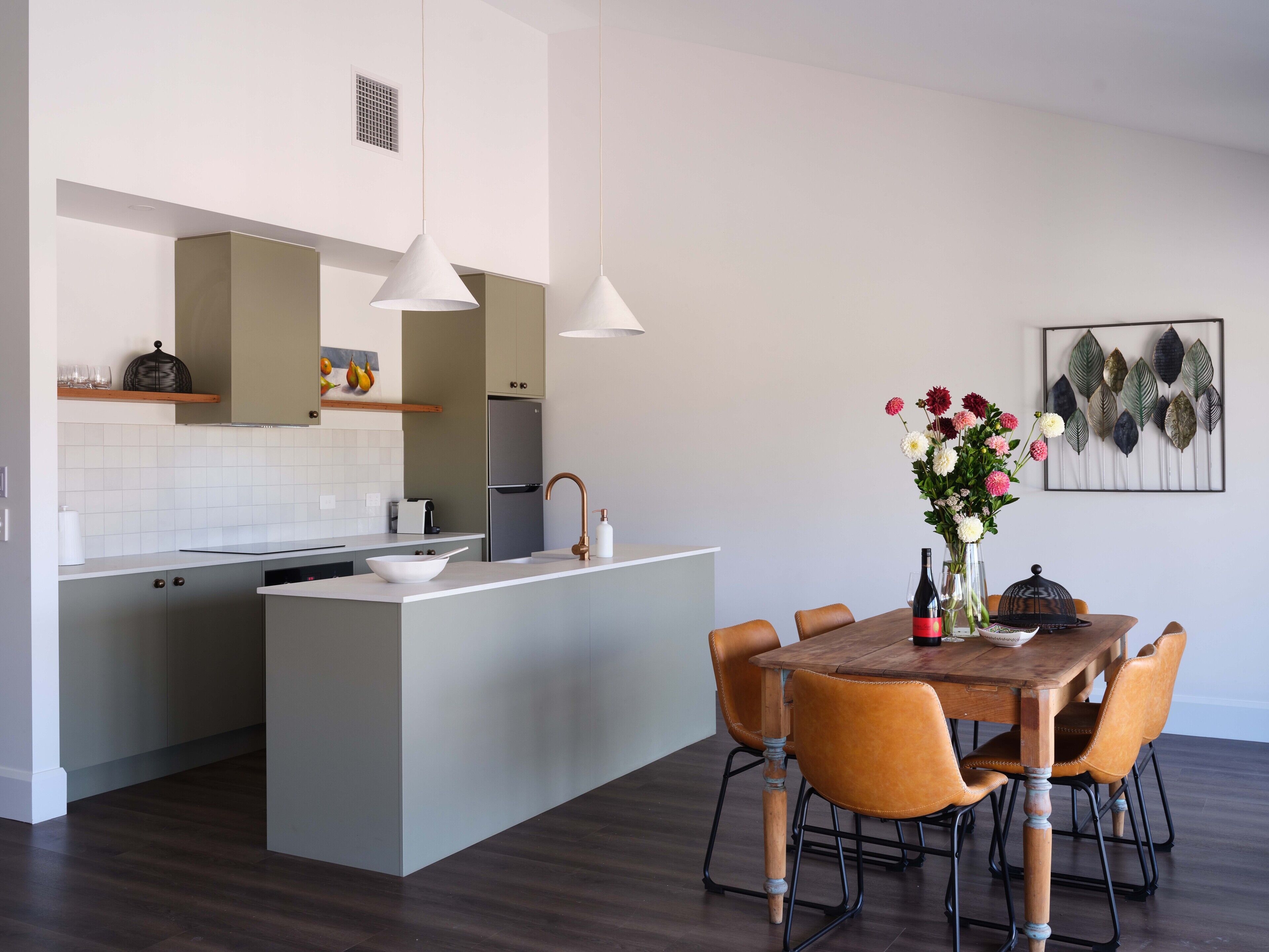 A modern kitchen with a central island, a dining area with a wooden table and chairs, and a decorative leaf wall art piece.