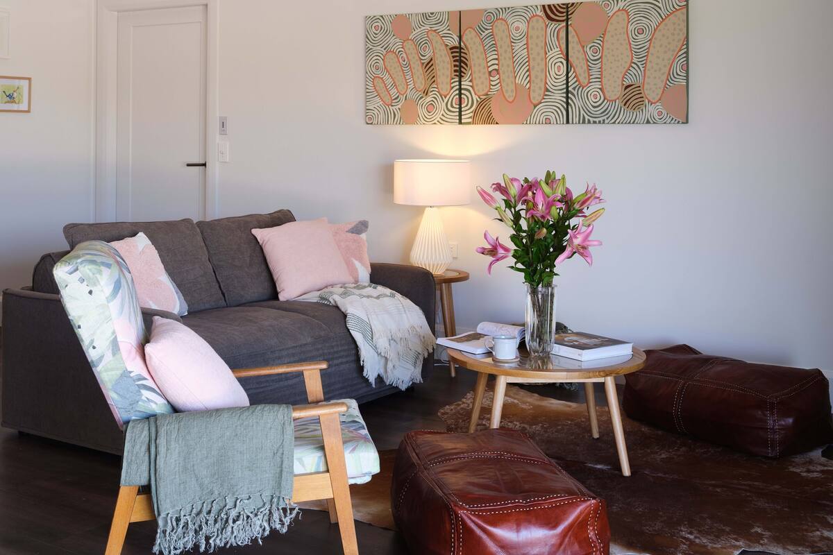 A living room with a grey sofa, a wooden chair, a small round table with a vase of flowers, a lamp, and a brown leather ottoman.
