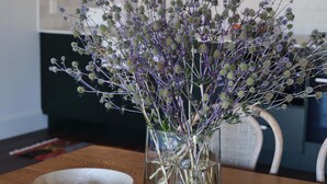 A dining table with a glass vase containing purple flowers, a wooden bowl with fruits, and a woven placemat. - Shawwood Estate (Bombira)