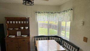 A kitchen with a wooden table and bench, a built-in shelf with dishes, and a window with a view of the backyard. - Cabin Near Sequoia & Kings Canyon National Parks (Dunlap)