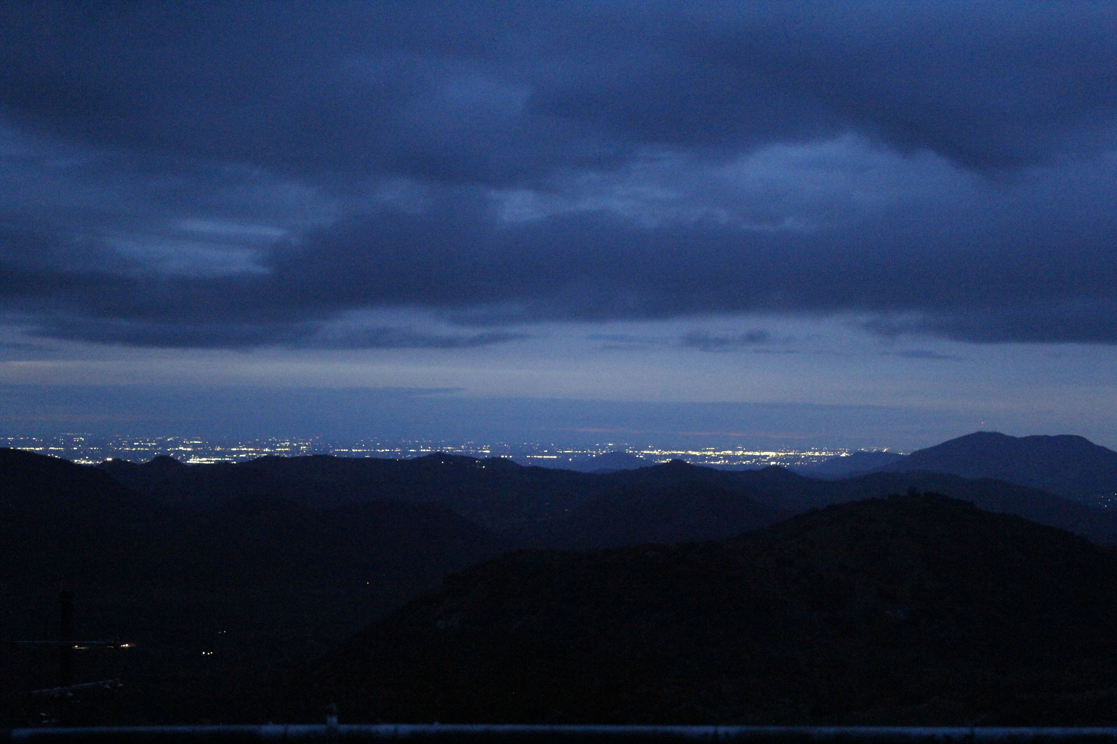 Ein Nachtblick auf eine Stadtlandschaft mit fernen Stadtlichtern und Bergsilhouetten unter einem bewölkten Himmel.