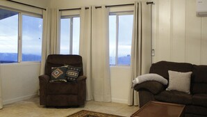 A living room with a brown armchair, a brown sofa, and a wooden coffee table. There are three windows with curtains, and a view of the mountains is visible through the windows. - Cabin Near Sequoia & Kings Canyon National Parks (Dunlap)