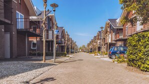 Une rue résidentielle bordée de maisons modernes, un ciel dégagé et une voiture bleue garée sur le côté droit.