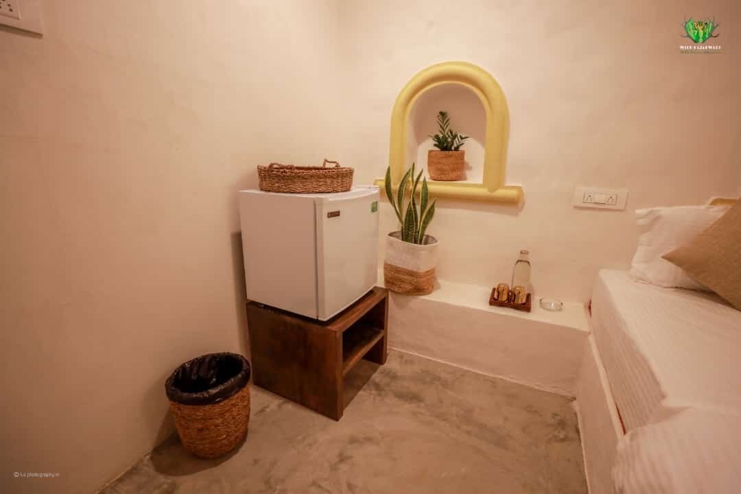 Una habitación de hotel pequeña con una cama, un refrigerador compacto, una planta en maceta, una cesta de mimbre y un cesto de basura.