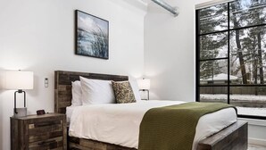 A bedroom with a wooden bed frame, white bedding, a green throw blanket, and a large window with a view of trees. - Rooftop Hot Tubs I Dogs Welcome I Near DT TC (Traverse City)
