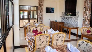 A dining area with multiple tables and chairs, a television mounted on the wall, and a window providing natural light. - Arusha Diplomat Hotel (Arusha)