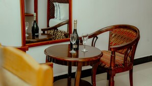A wooden dining table with a bottle and two glasses, a wooden chair with a patterned cushion, and a mirror reflecting a bed and a patterned wall. - Arusha Diplomat Hotel (Arusha)