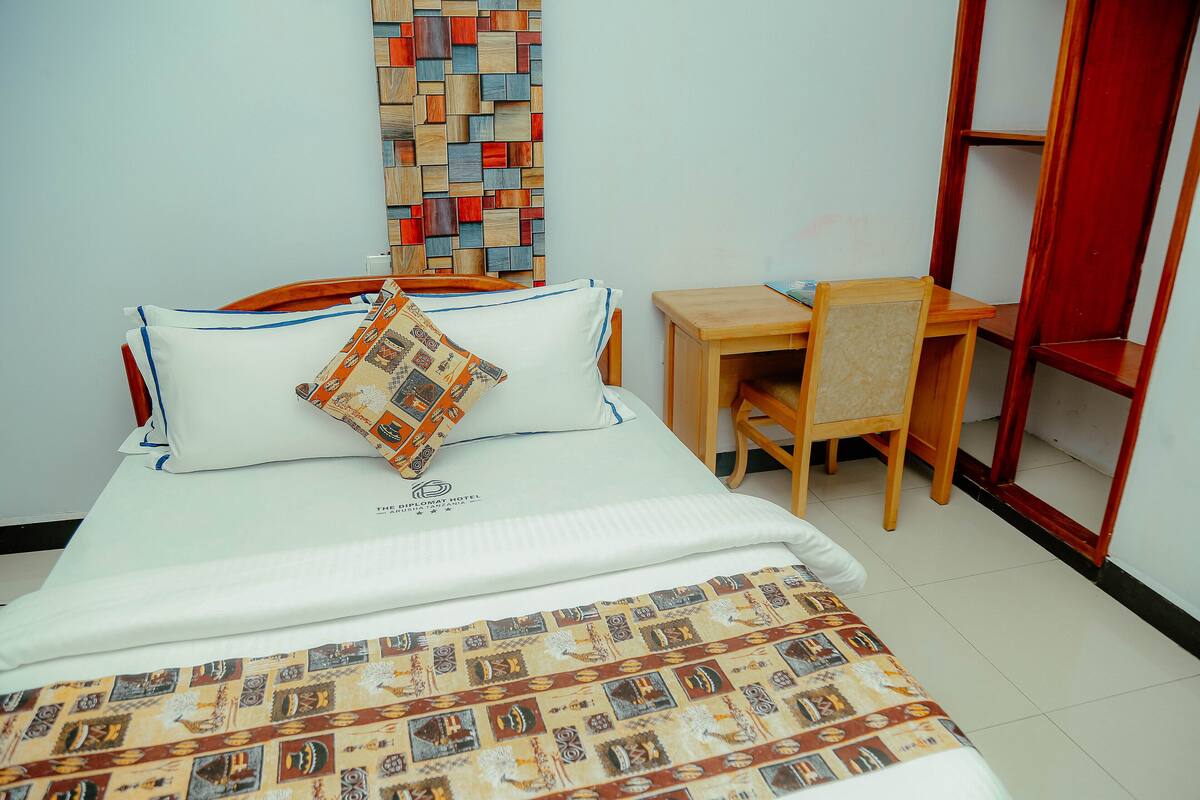 A neatly made bed with a patterned bedspread, a pillow, and a matching pillow on the headboard. A small wooden desk with a chair is positioned beside the bed.