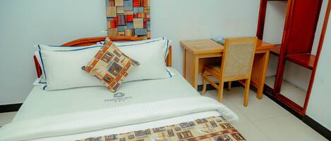 A neatly made bed with a patterned bedspread, a pillow, and a matching pillow on the headboard. A small wooden desk with a chair is positioned beside the bed.