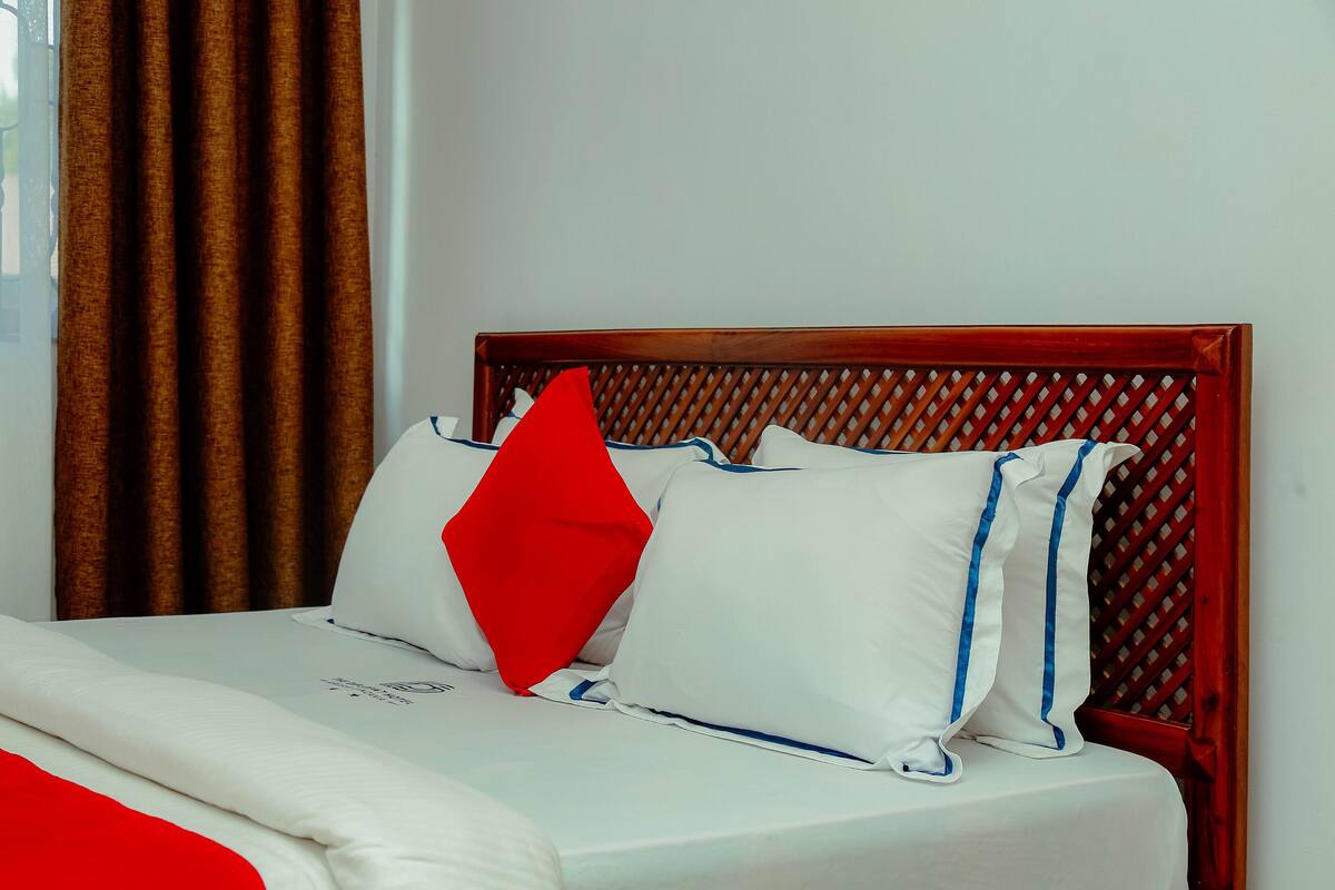 A neatly made bed with white bedding and a red pillow, a wooden headboard, and brown curtains.