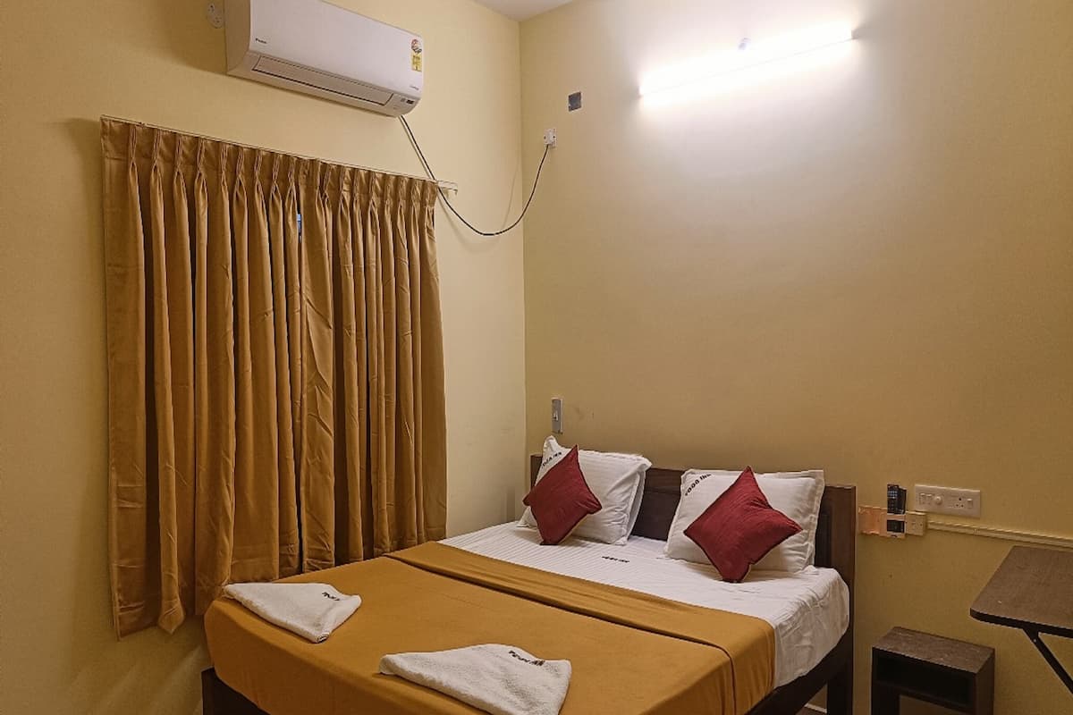 A hotel room with a bed, a small table, a chair, an air conditioning unit, and a window with curtains.