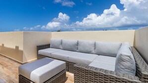 Una terraza en la azotea con un sofá y reposapiés de mimbre, un cielo azul despejado y un paisaje arenoso al fondo.