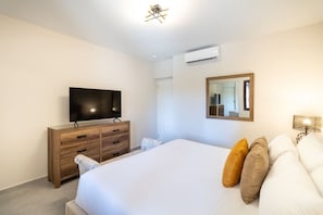 A hotel room with a bed, a TV on a wooden dresser, an air conditioner, a mirror, and a bedside lamp.