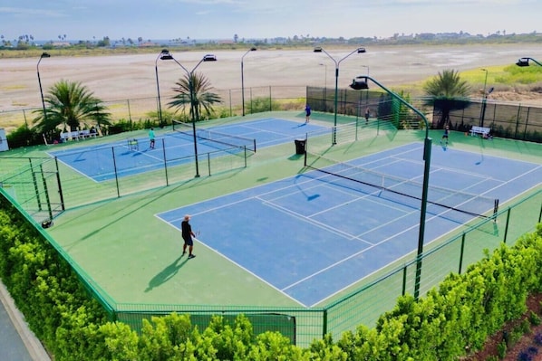 A tennis court with a net, surrounded by a green fence, and palm trees in the background.