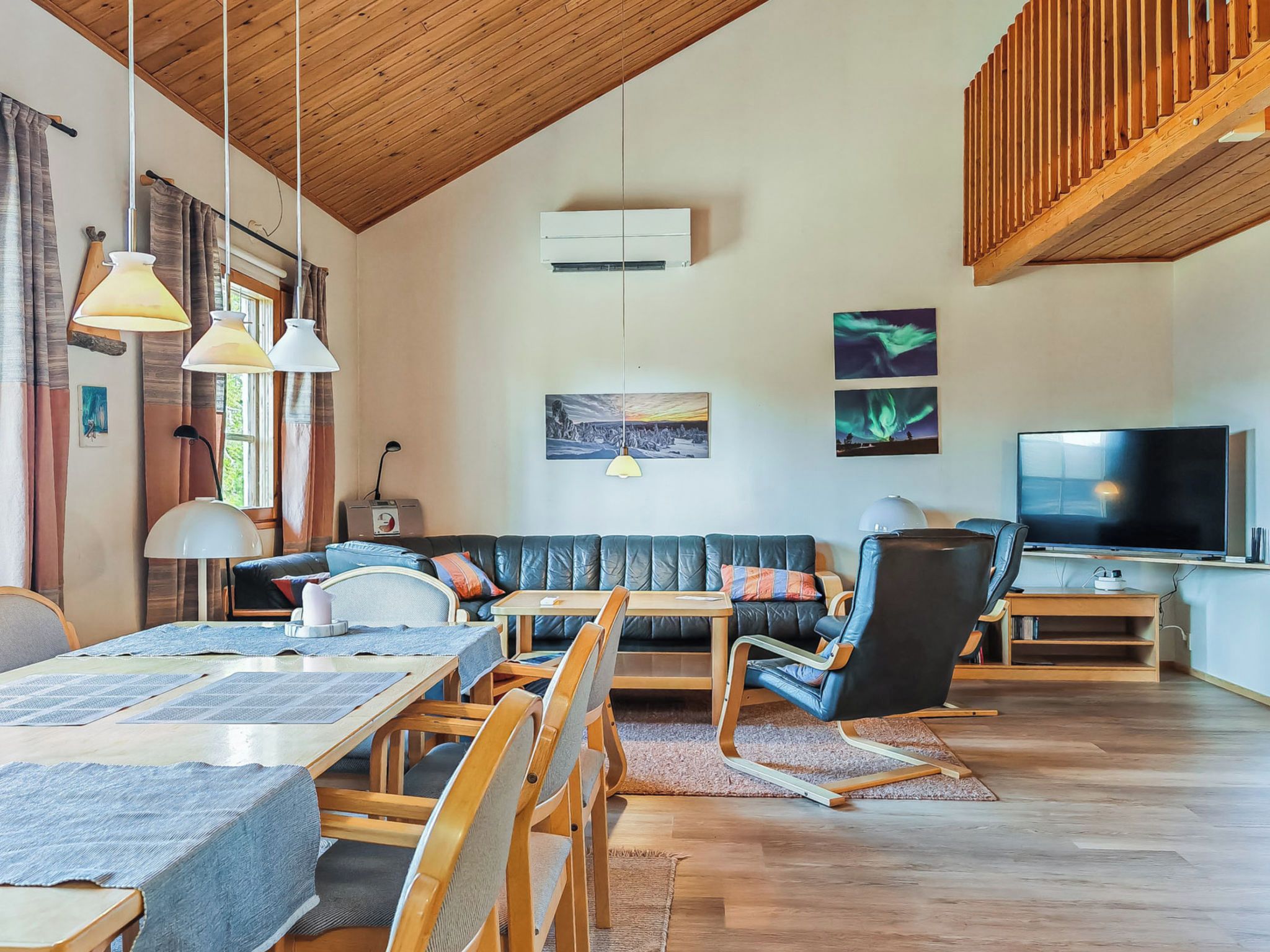 Un salon moderne avec un plafond en bois, un canapé en cuir noir, une table à manger avec des chaises, un téléviseur et des œuvres d’art accrochées aux murs.