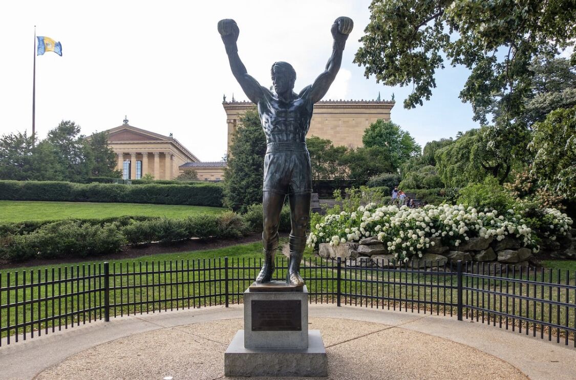 A statue of a boxer with raised arms in a park setting, surrounded by a fence and greenery.
