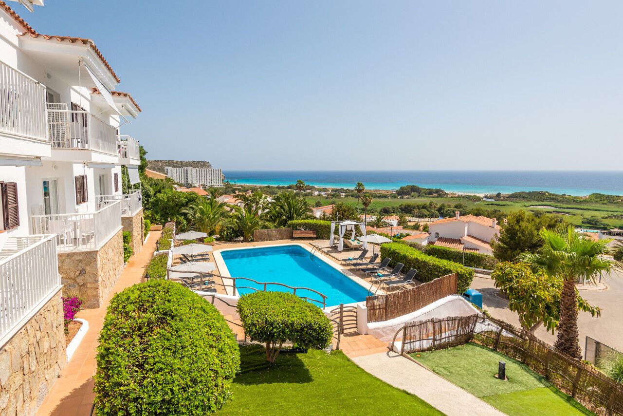 A pool area with a clear view of the ocean, surrounded by well-maintained lawns and a white building with balconies.