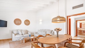 A modern living room with a wooden dining table, wicker chairs, a sofa set, and a TV mounted on the wall.