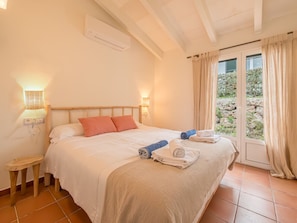Quarto com cama, abajures, uma mesinha e vista através de uma porta de vidro.