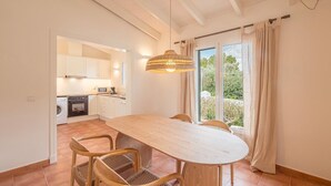 Un'ampia sala da pranzo con un lungo tavolo in legno e sedie in vimini, una cucina sullo sfondo e un'ampia finestra con tende.