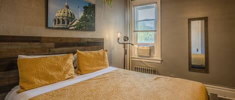 Une chambre à coucher avec un grand lit, un tableau représentant un bâtiment accroché au mur, une fenêtre avec des stores et un radiateur.
