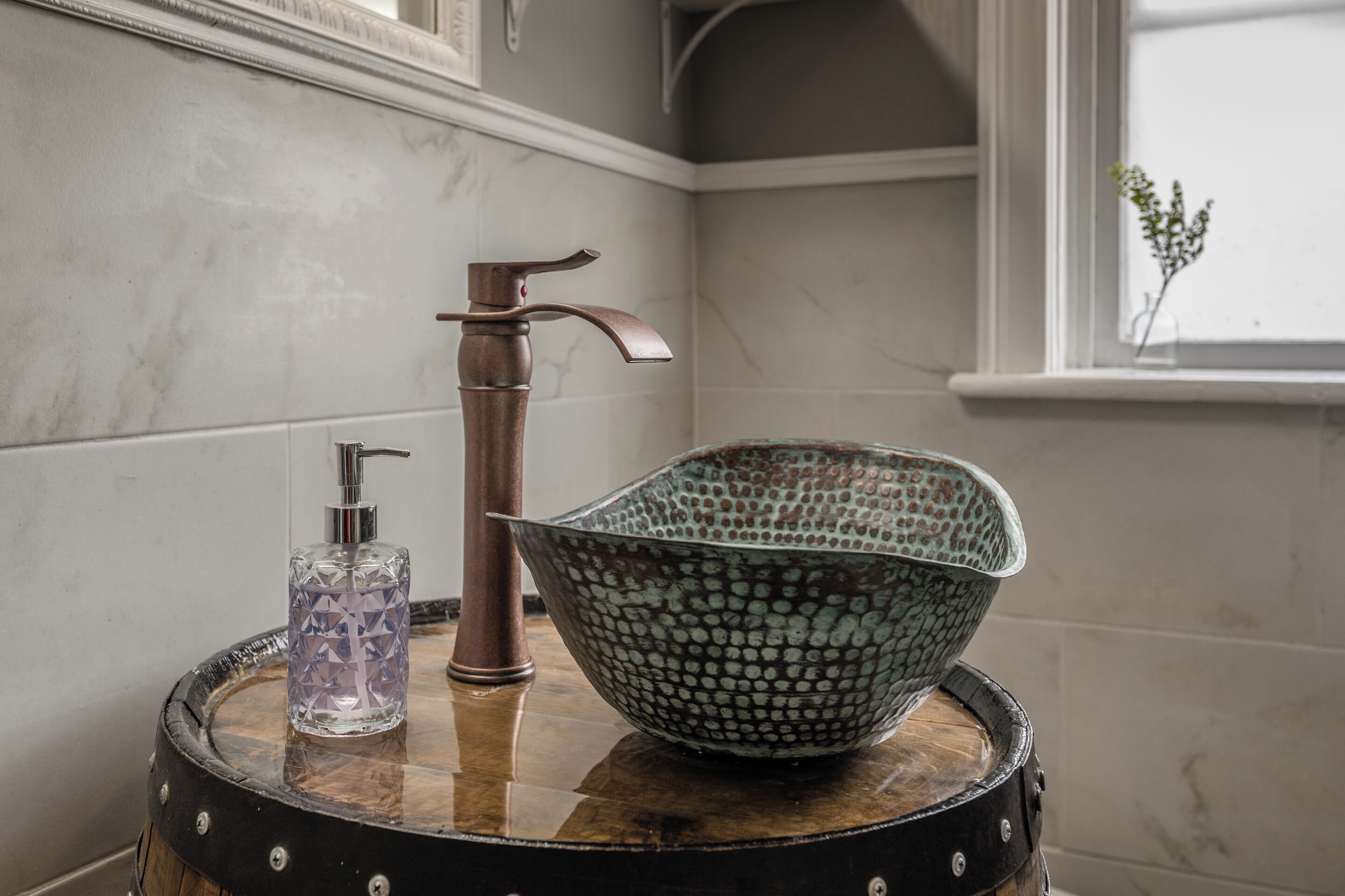 A bathroom with a wooden barrel stool, a glass bottle, and a metal faucet.
