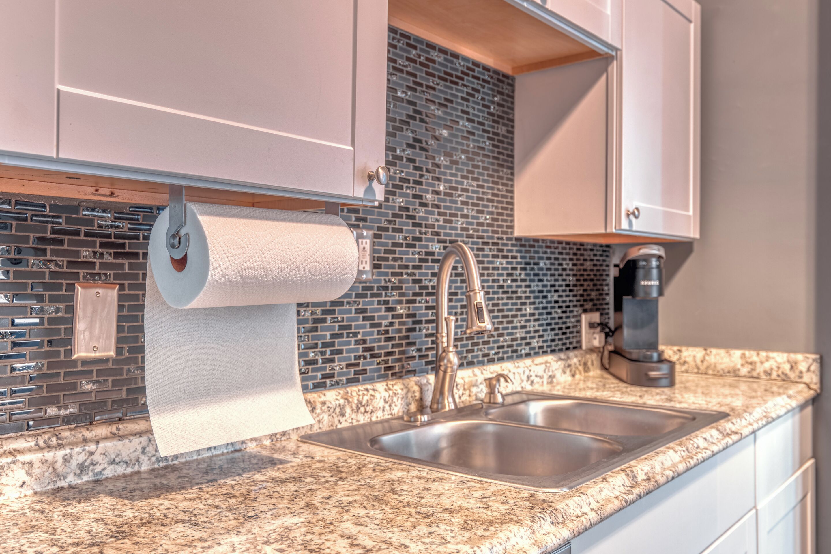 A kitchen with a granite countertop, stainless steel sink, and a roll of paper towels hanging from the cabinet.