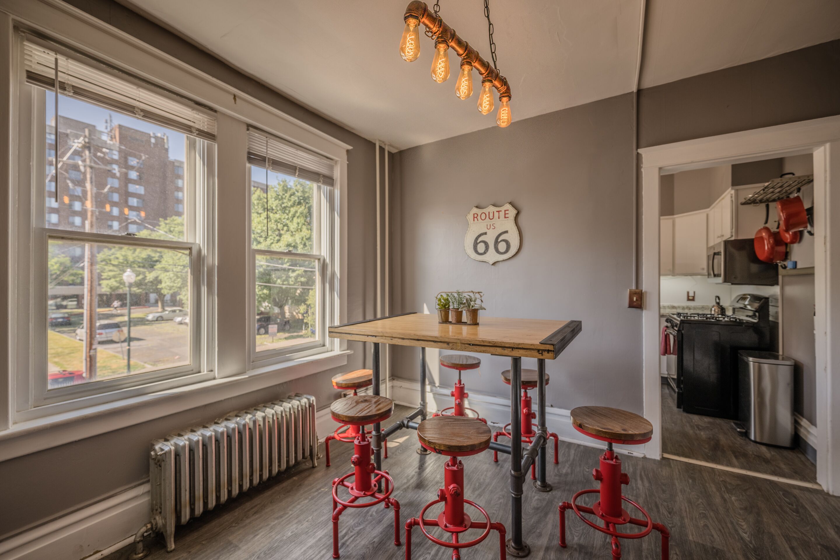A kitchen with a wooden table and red stools, a window with a view of buildings, and a sign on the wall reading "Route 66".
