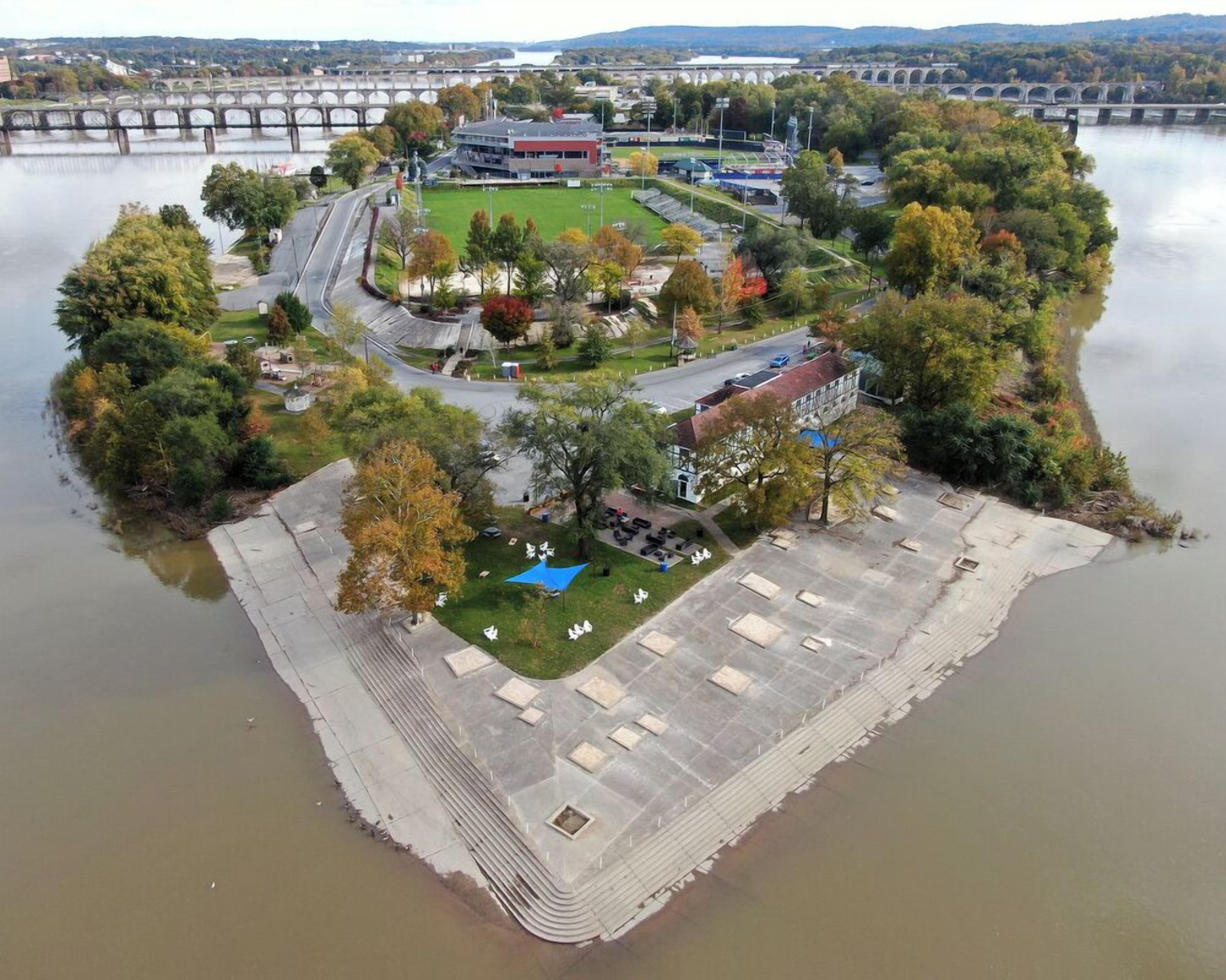 Luftaufnahme eines Flussuferparks mit einer Brücke, Gebäuden und einem Parkplatz.
