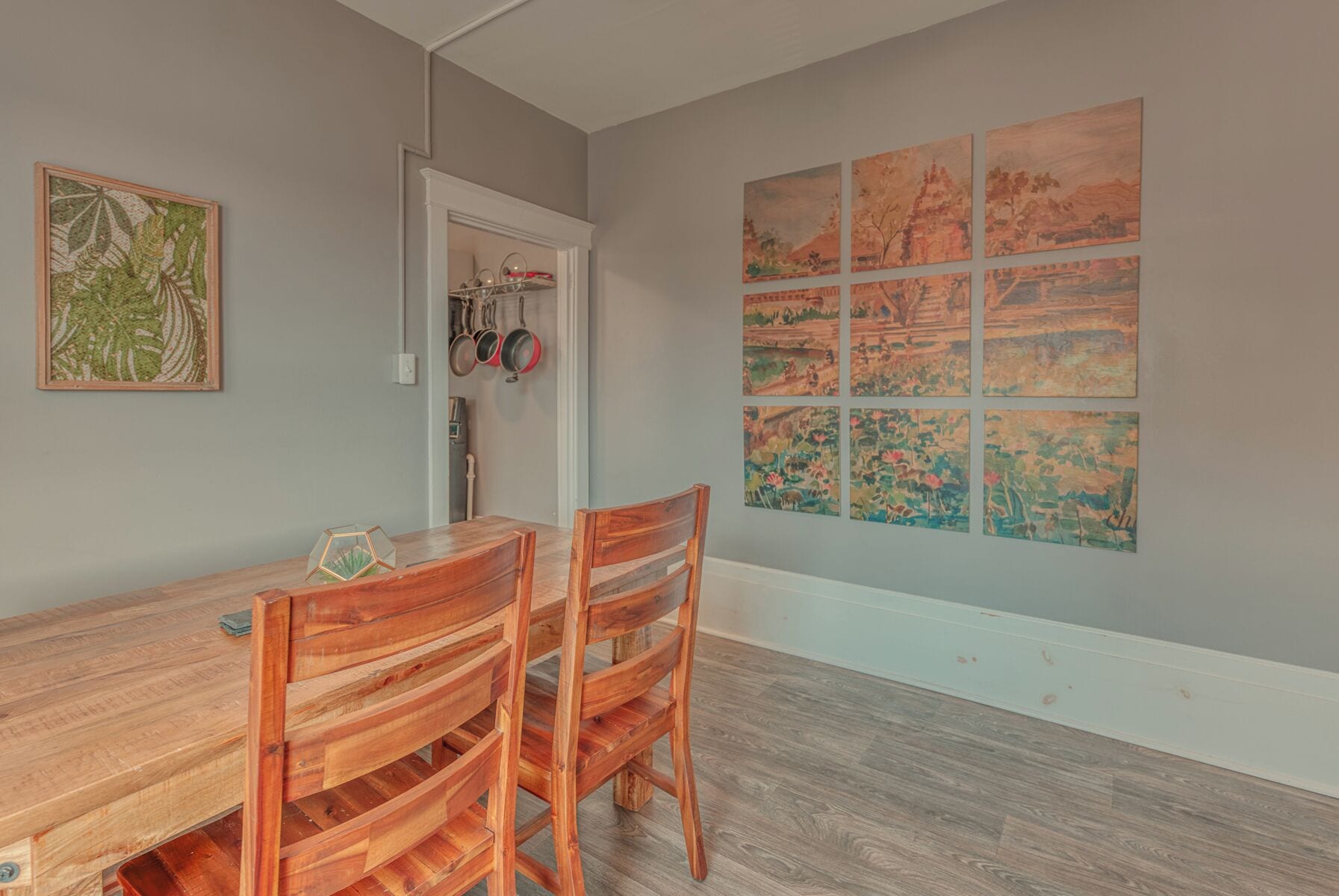 A dining area with a wooden table and chairs, a framed botanical print, and a wall with multiple landscape paintings.