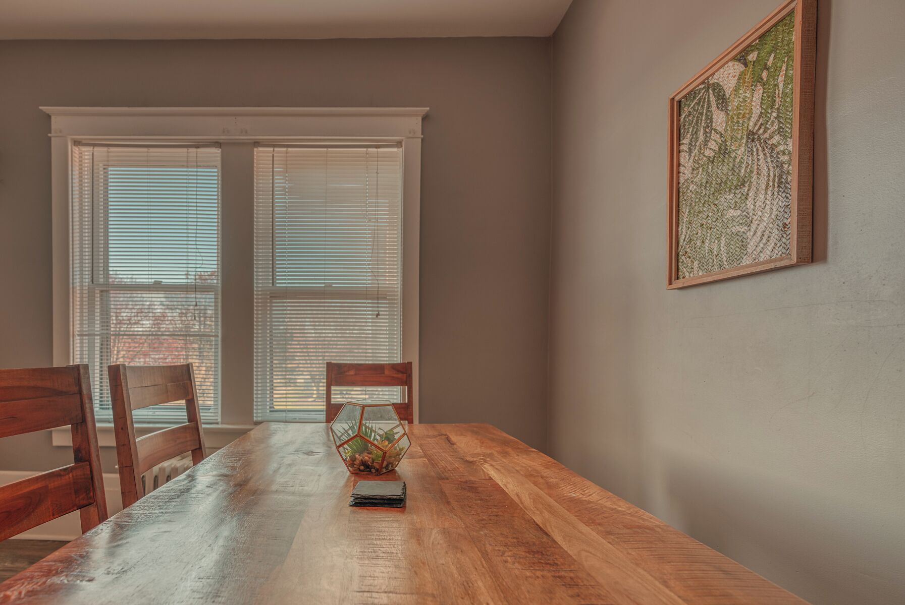 A dining area with a wooden table, two chairs, a hexagonal glass-top table lamp, and a framed artwork on the wall.