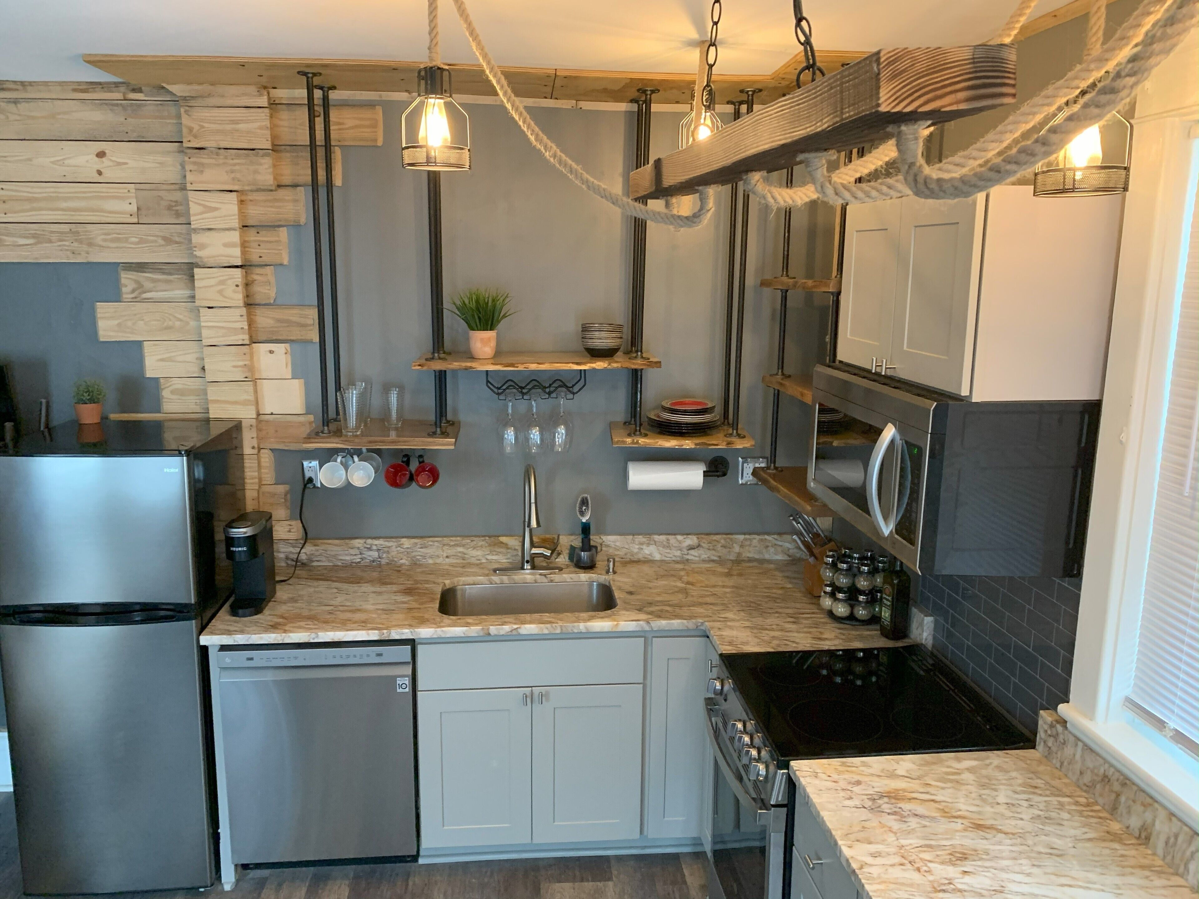 A modern kitchen with a stainless steel refrigerator, dishwasher, and stove. There are wooden shelves with plants and kitchenware, and a hanging light fixture.